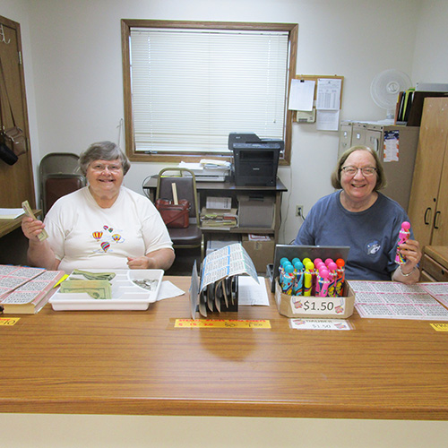 Two women sitting at a desk selling bingo cards and markers